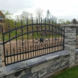 Czarne, kute ogrodzenie z kamiennymi słupkami, widok na budynek gospodarczy i silosy w tle.