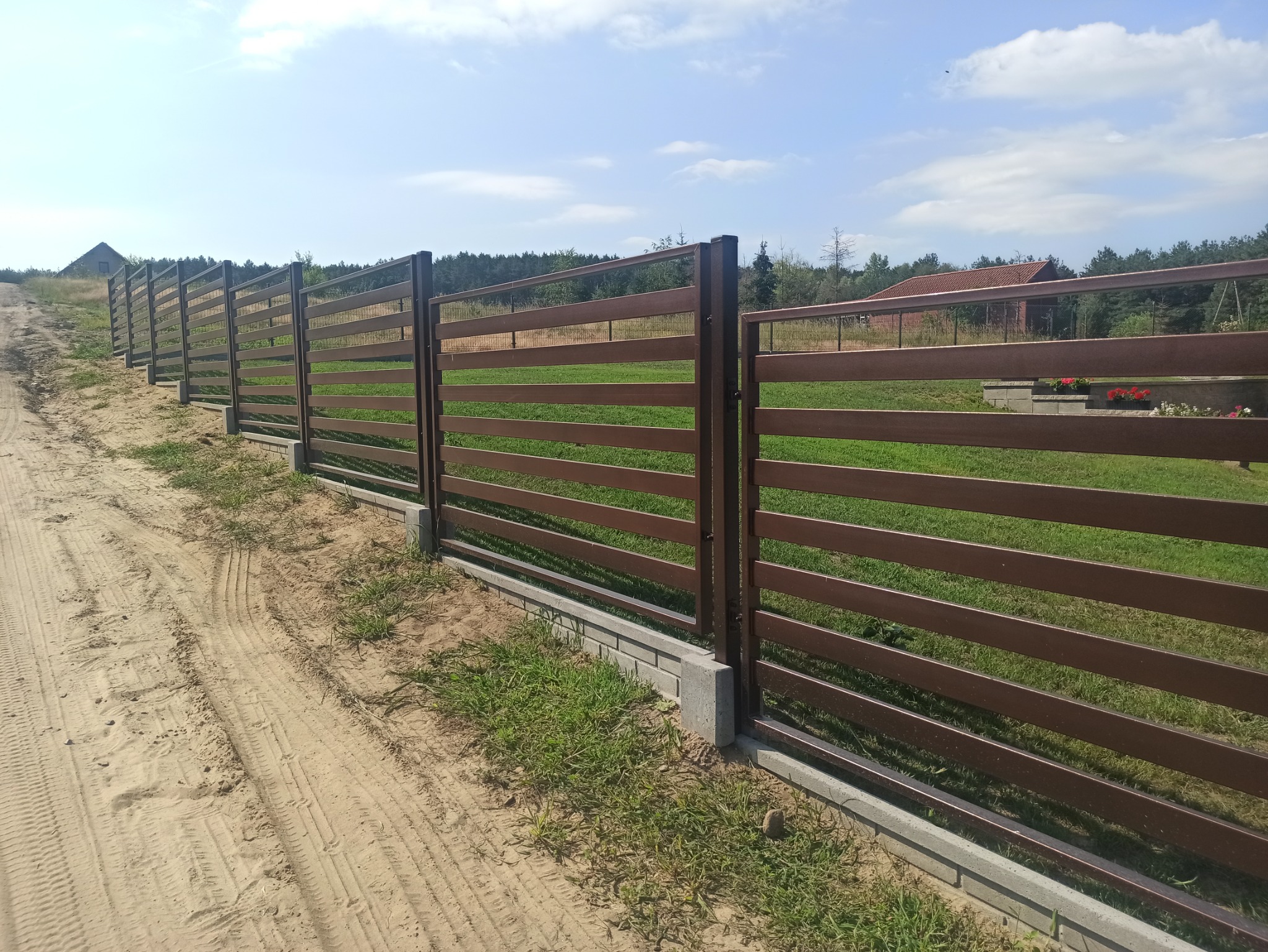 Długi, horyzontalny płot z brązowych, poziomych desek i metalowych słupków na betonowej podmurówce, widok z boku na piaszczystej drodze, w tle zieleń i budynki.
