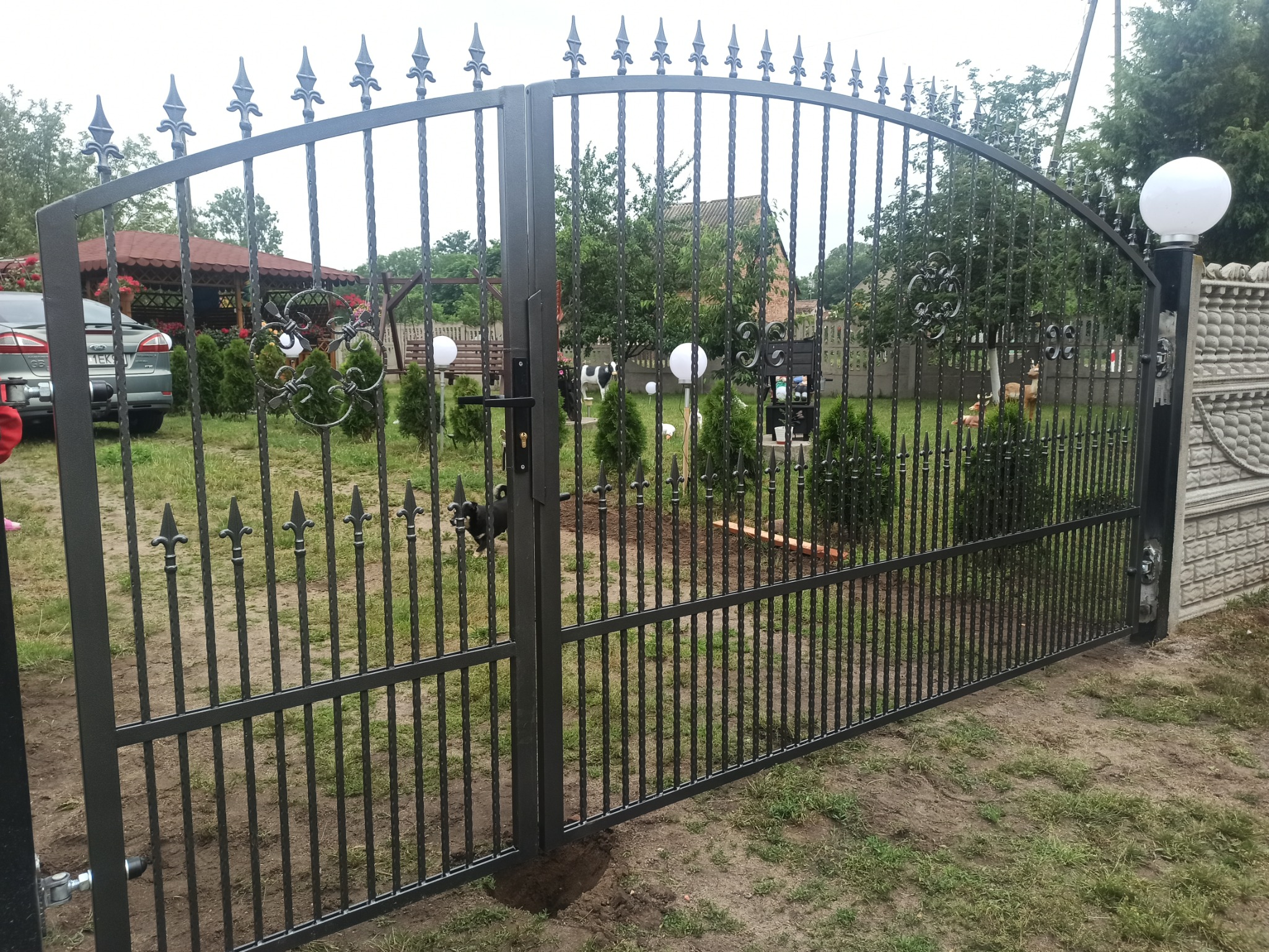 Nowoczesna, dwuskrzydłowa brama wjazdowa z metalowymi prętami i dekoracyjnymi elementami, widok z zewnątrz na posesję z ogrodem i budynkami.