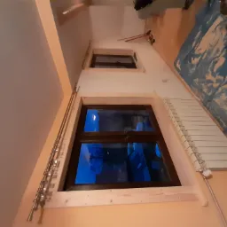 Montaż okna dachowego z drewnianą ramą, widoczne narzędzia i zabezpieczenia na podłodze, grzejnik obok okna, wieczorny widok za oknem.