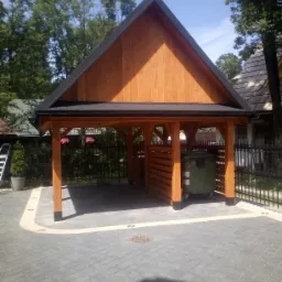 Drewniana wiata ze skośnym dachem kryjąca pojemnik na śmieci, widoczna na tle brukowanej powierzchni i ogrodzenia.