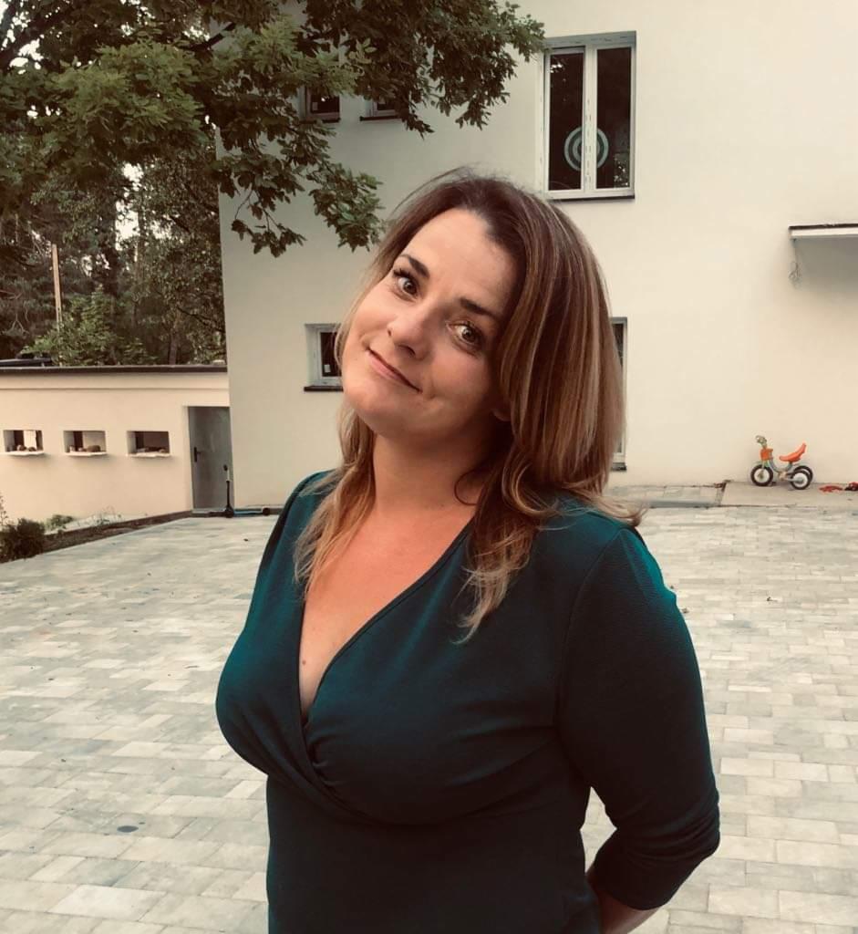 Uśmiechnięta kobieta w zielonej sukience na tle jasnego budynku z oknami i kostki brukowej. W tle widoczny rowerek dziecięcy.