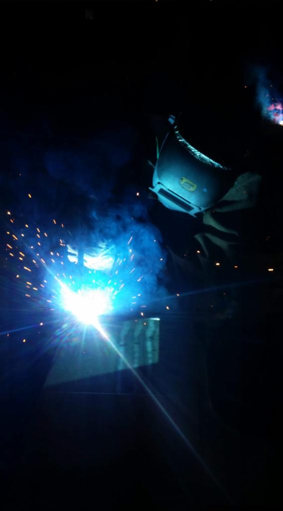 Spawanie metalu w ciemnym otoczeniu, widoczne iskry i niebieski dym, spawacz w masce ochronnej.