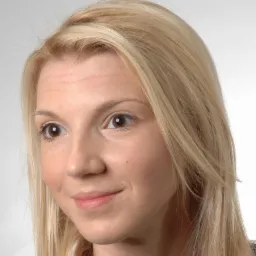 Portret uśmiechniętej kobiety o jasnych włosach, z delikatnym makijażem, patrzącej w bok na jasnym tle. Widoczny pieprzyk na policzku.