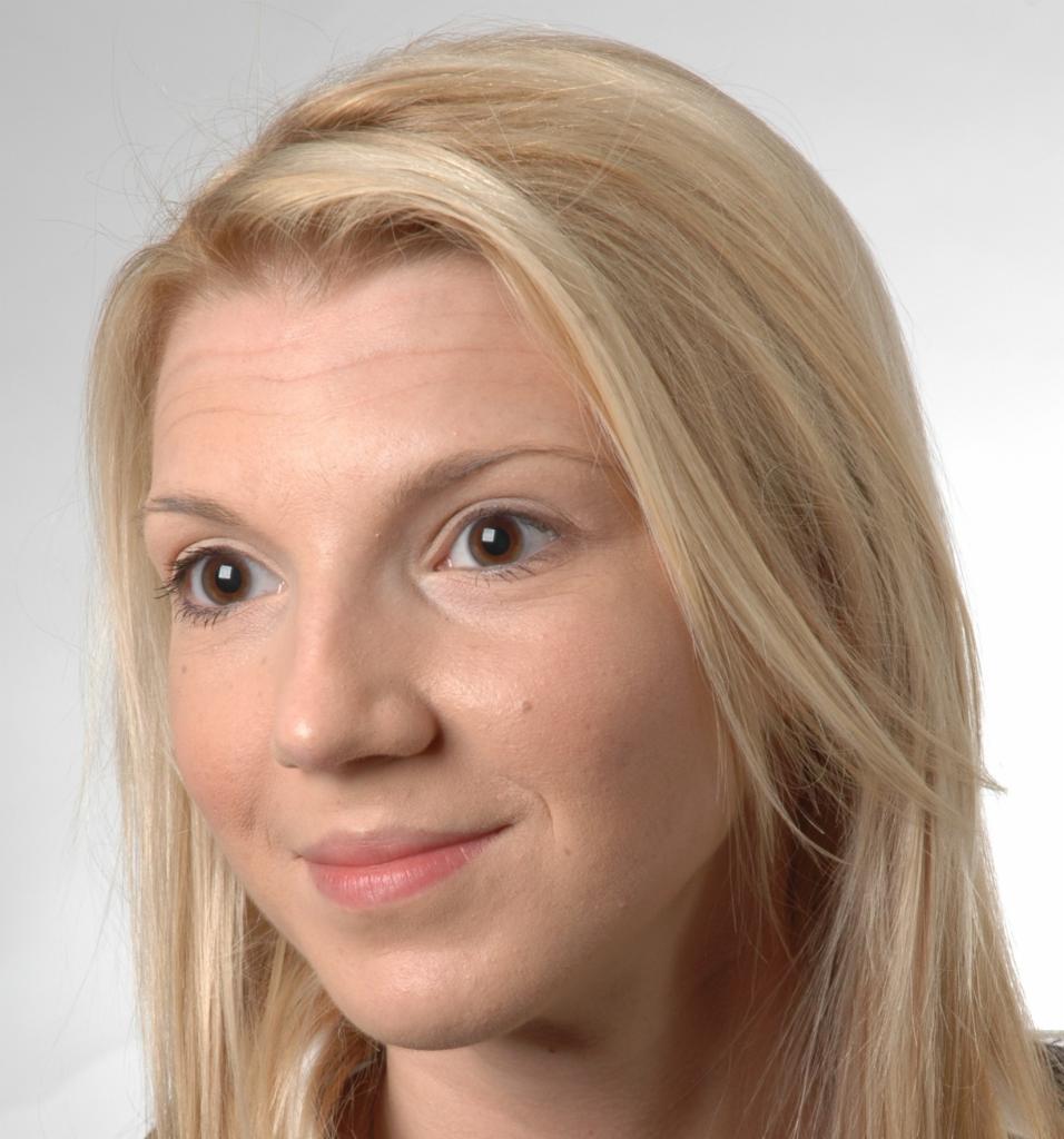 Portret uśmiechniętej kobiety o jasnych włosach, z delikatnym makijażem, patrzącej w bok na jasnym tle. Widoczny pieprzyk na policzku.