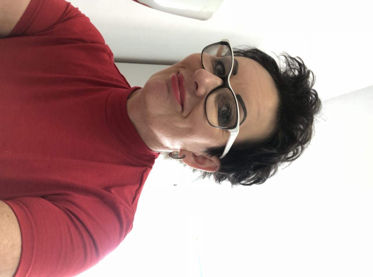 Portret kobiety w czerwonej koszulce i okularach z białymi oprawkami, uśmiechająca się do kamery, widok z dołu.