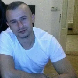 Portret uśmiechniętego mężczyzny w białej koszulce, z kr&oacute;tką fryzurą, siedzącego w jasnym wnętrzu. Widoczny fragment pomieszczenia z drzwiami.