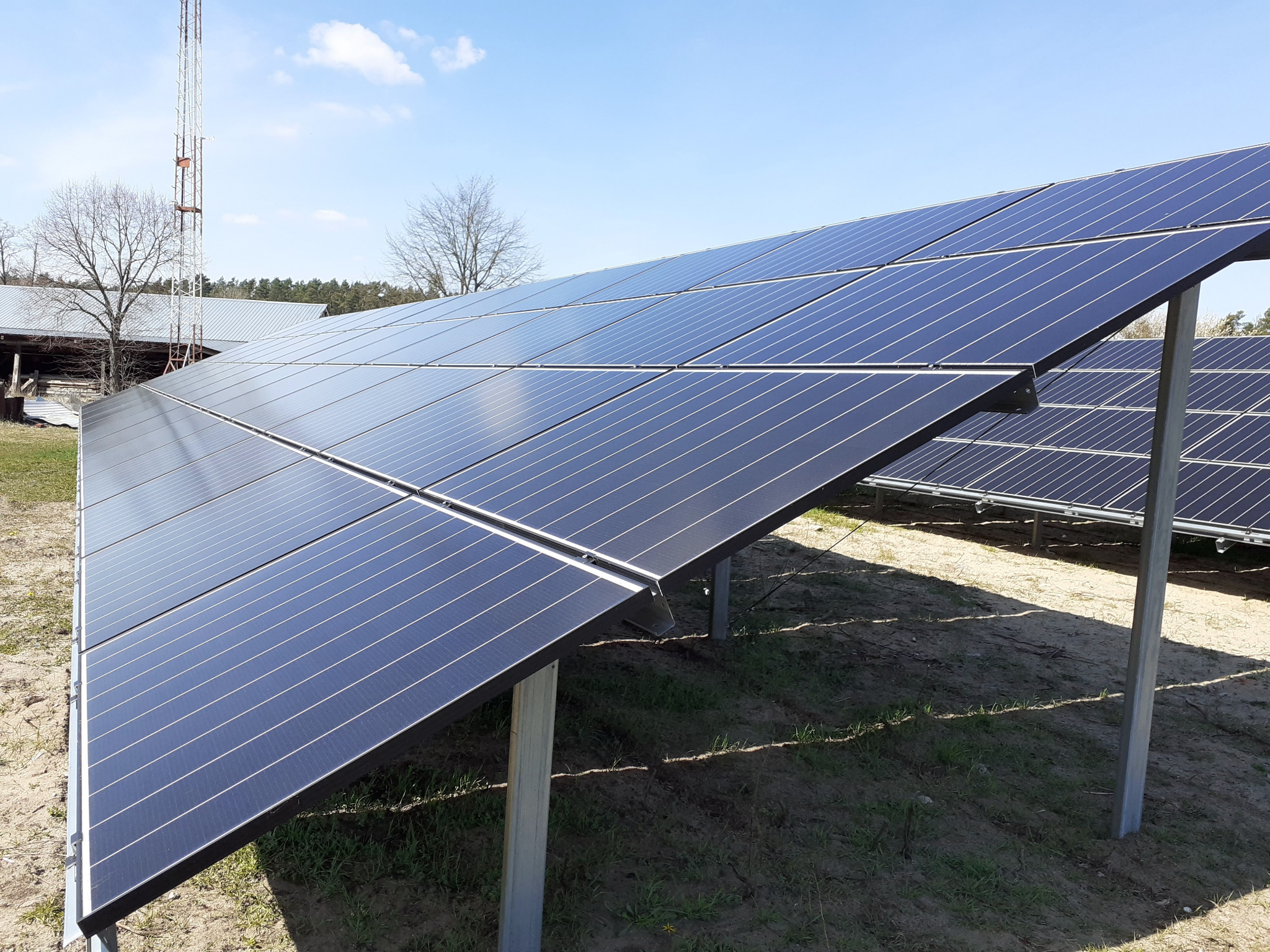 Rząd paneli słonecznych ustawionych pod kątem na metalowych wspornikach, z widoczną wieżą telekomunikacyjną i budynkiem gospodarczym w tle, przy słonecznej pogodzie.