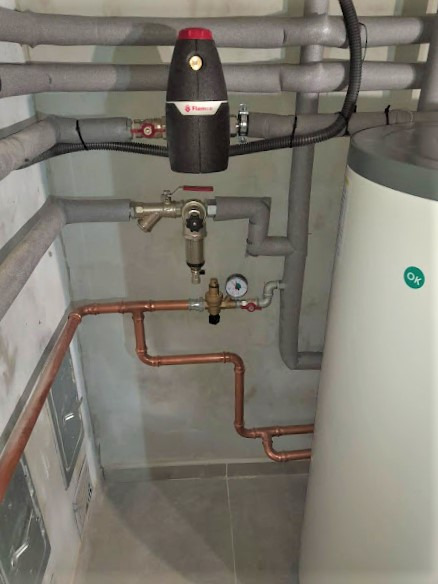 Instalacja grzewcza z pompą ciepła: widoczne rury miedziane, zbiornik akumulacyjny z zielonym logo OK, czarny moduł Flamco i zaizolowane rury szare.