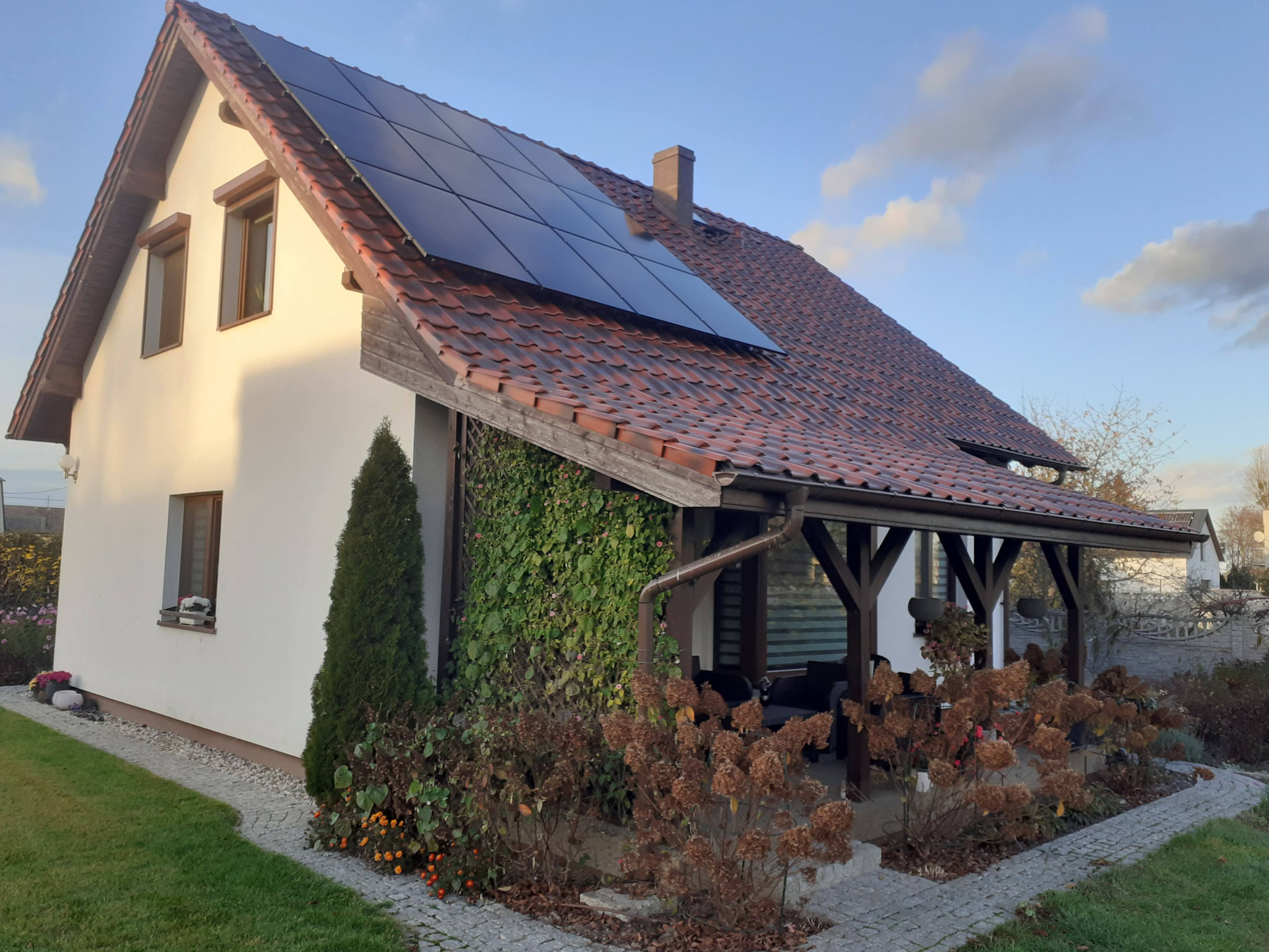 Dom jednorodzinny z czerwonym dachem i panelami słonecznymi na połaci dachu, z drewnianą altaną porośniętą bluszczem z boku.