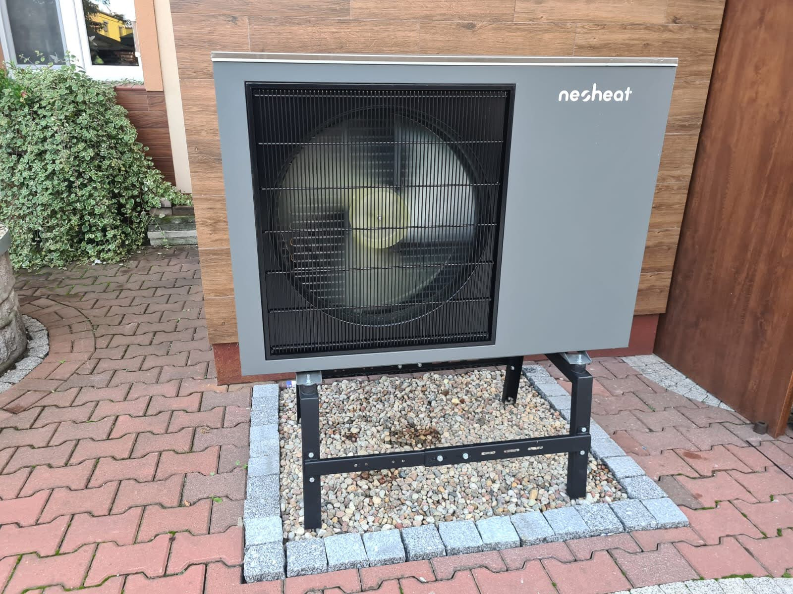 Szara pompa ciepła marki Nesheat zamontowana na czarnej, metalowej konstrukcji, otoczona ozdobnym obramowaniem z kostki brukowej i kamyków, na tle drewnianej elewacji domu.