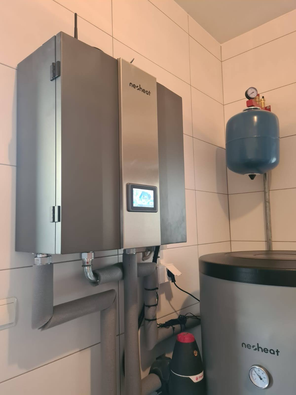 Szara pompa ciepła marki Nesheat zamontowana na kafelkowanej ścianie, widoczny ekran dotykowy, rury izolowane oraz zbiornik wyrównawczy i zasobnik ciepłej wody.