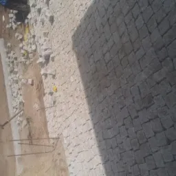 Układanie kostki brukowej na posesji, widoczne worki z materiałem, narzędzia i częściowo ułożona nawierzchnia. Praca w toku, ujęcie z góry.