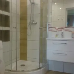 Nowoczesna kabina prysznicowa z zaokrąglonymi szybami, białe i beżowe płytki na ścianach, drewnopodobna podłoga, biała szafka z umywalką i lustro z oświetleniem w tle.