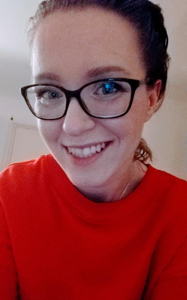 Portret uśmiechniętej kobiety w okularach i czerwonej bluzce, z włosami upiętymi w kok. Ujęcie z dołu, widoczne zęby i odbicie światła w szkłach.