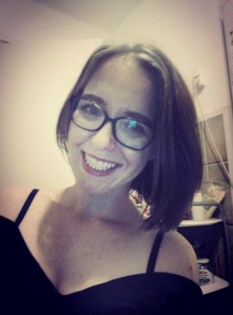 Uśmiechnięta kobieta w okularach z krótkimi brązowymi włosami, ubrana w czarny strój z ramiączkami, pozująca w pomieszczeniu z półkami w tle.