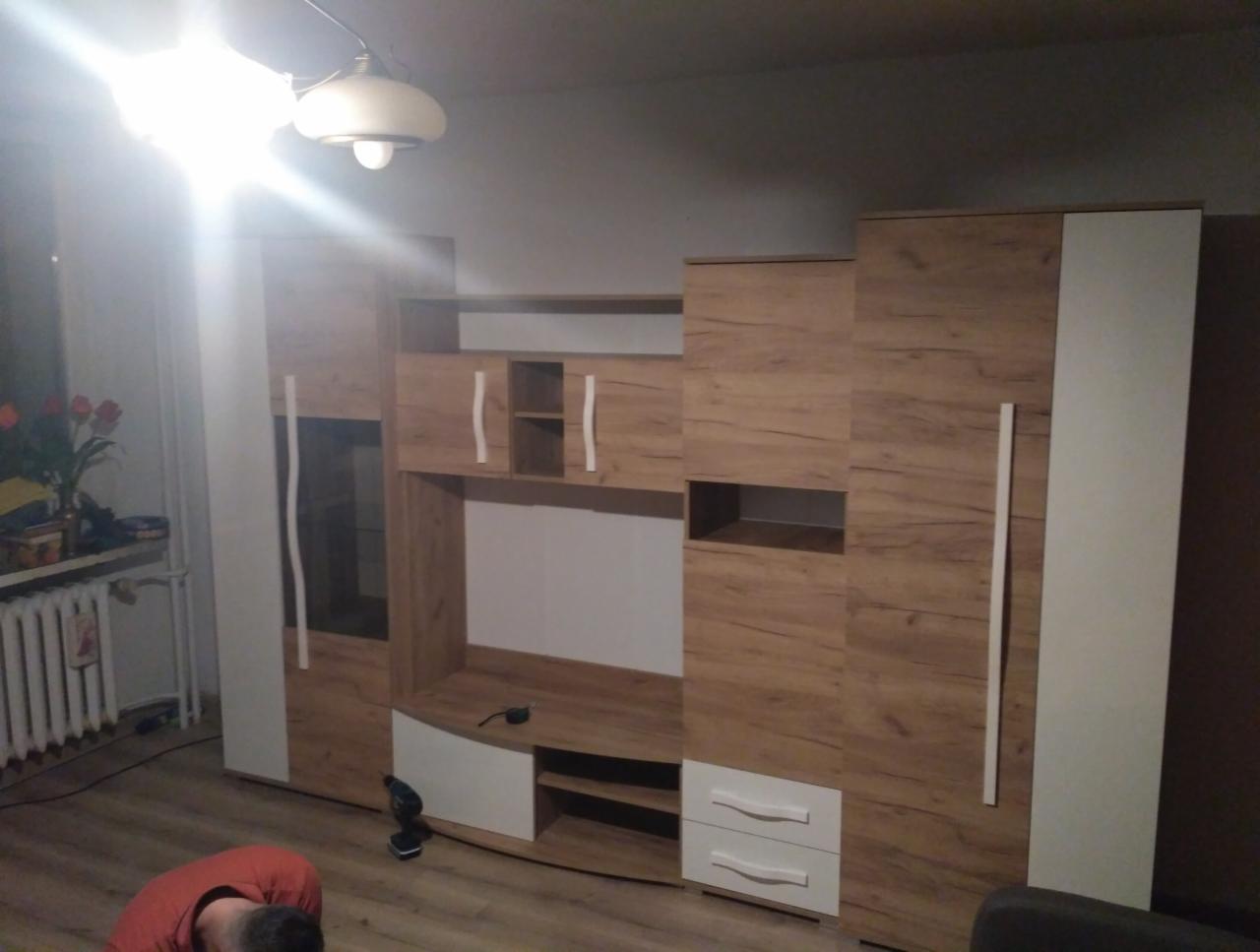 Zmontowany zestaw mebli do salonu z frontami w odcieniach drewna i białymi akcentami, widoczny fragment osoby w czerwonej koszulce oraz lampa sufitowa