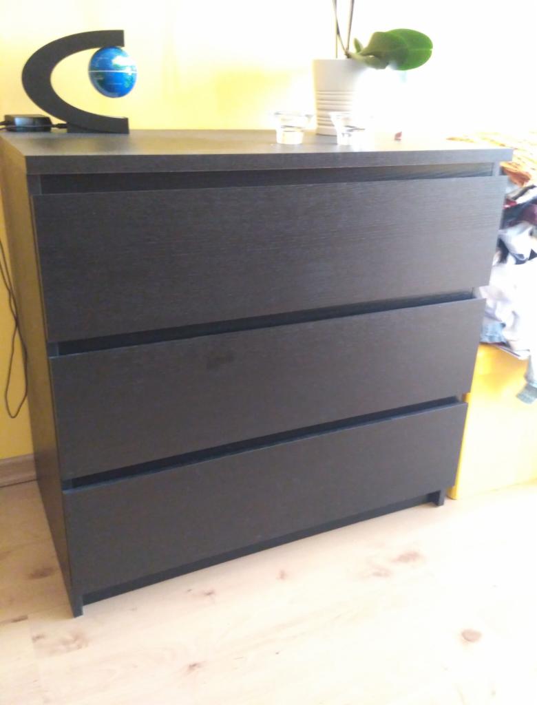 Ciemna, drewnopodobna komoda z trzema szufladami, na blacie dekoracje: globus w czarnej oprawie i biała doniczka z rośliną, widoczne niedoskonałości w wykończeniu.
