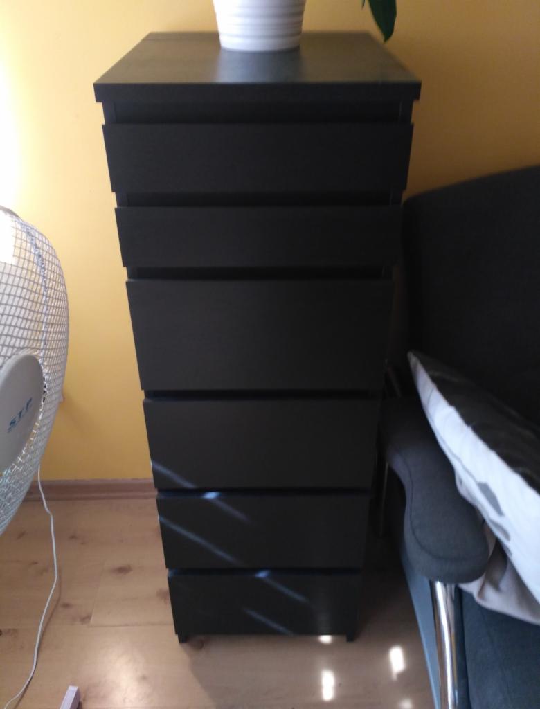 Wysoka, czarna komoda z sześcioma szufladami, na której stoi biała doniczka z rośliną, obok szarego fotela z poduszką, na drewnianej podłodze i żółtej ścianie w tle.