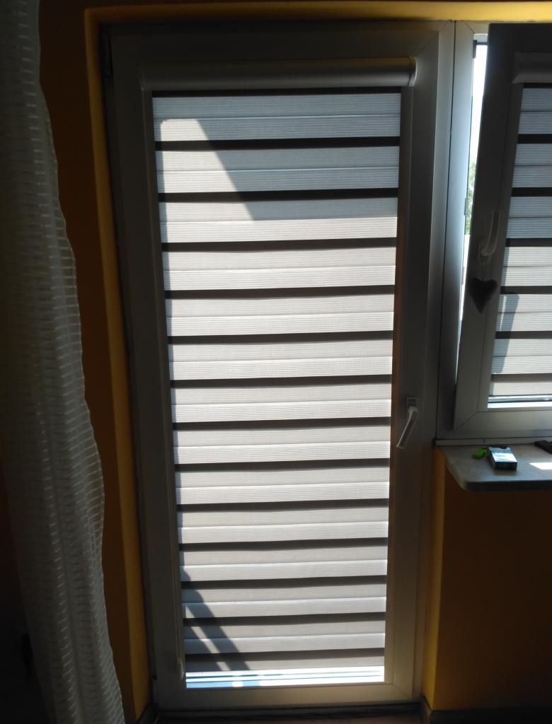 Okno z zamontowanymi roletami dzień-noc w kolorze szaro-białym, z widocznym fragmentem firanki po lewej stronie i parapetem z telefonem po prawej.