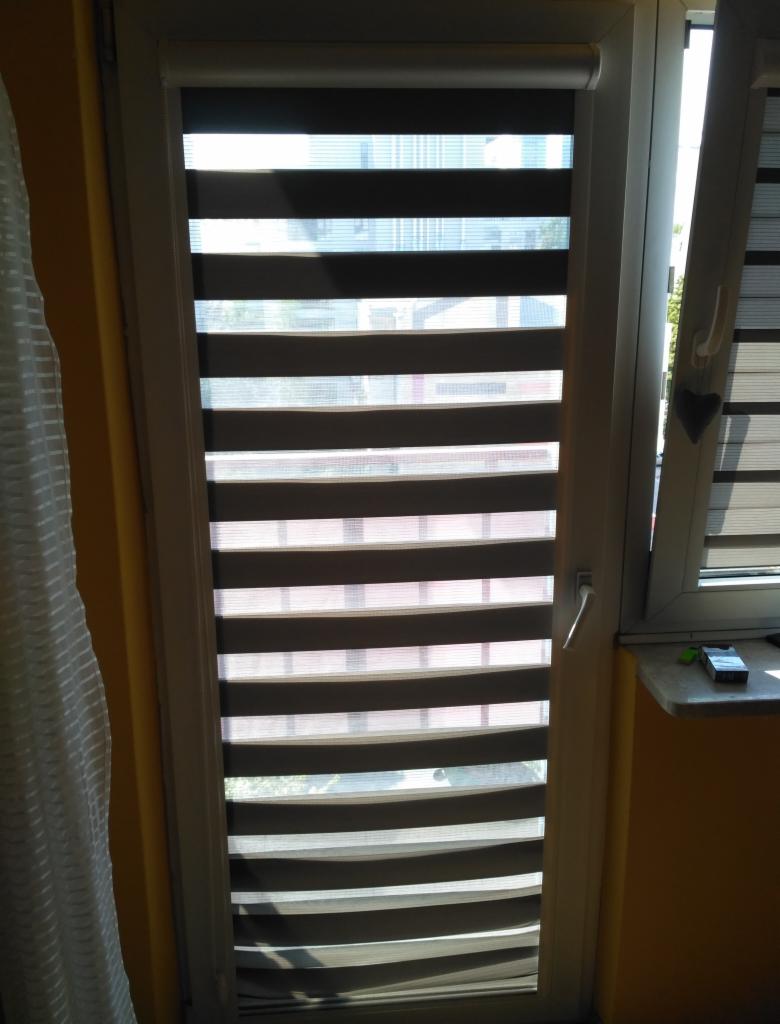 Okno z roletą dzień-noc w kolorze szaro-białym, zamontowaną w białej ramie okiennej, obok fragment białej zasłony i parapetu z drobnymi przedmiotami.