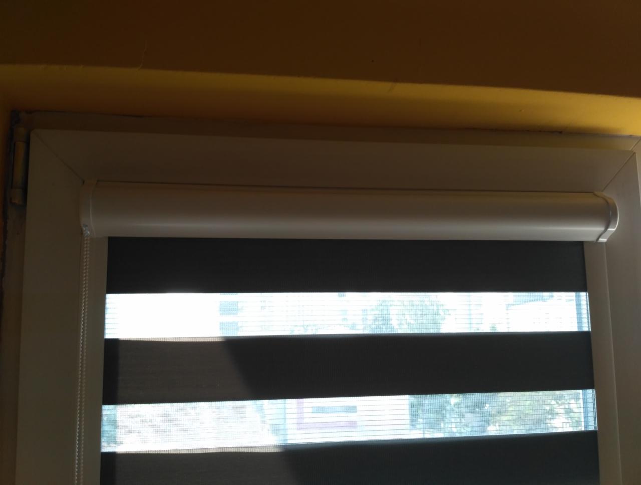 Okno z roletą zebra w kolorze szaro-białym, widoczne z bliska, z żółtą ścianą w tle u góry.