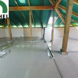 Świeżo wylana, jasna posadzka w surowym wnętrzu poddasza z widoczną drewnianą konstrukcją dachu i logo firmy KOCHEL w lewym górnym rogu.