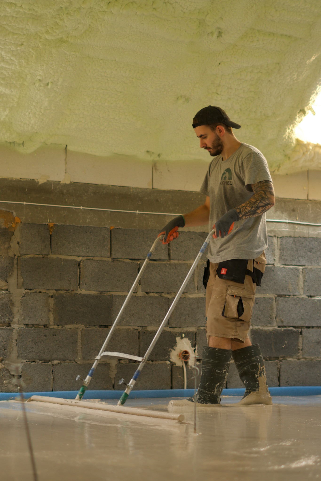 Pracownik w kaloszach i roboczych spodniach rozprowadza płynny materiał na posadzce za pomocą specjalistycznego narzędzia z długim uchwytem; ściany z bloczków, sufit ocieplony pianą.