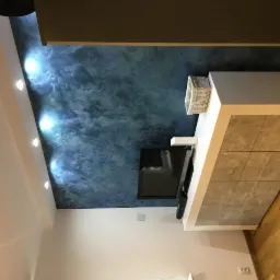 Ściana w salonie z efektem dekoracyjnym imitującym marmur w odcieniach granatu i błękitu, oświetlona punktowymi lampami sufitowymi, z telewizorem i białą szafką RTV z szarymi frontami.