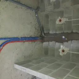 Surowy stan łazienki w trakcie remontu: widoczne rury ciepłej i zimnej wody w izolacji, szare rury kanalizacyjne z trójnikiem, bruzdy w ścianie z instalacją elektryczną i fragment wykafelkowanej...