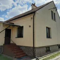 Jasnobeżowy dom jednorodzinny z brązowymi oknami, rynnami i podmurówką z cegły, numer domu 79, błękitne niebo z chmurami w tle.