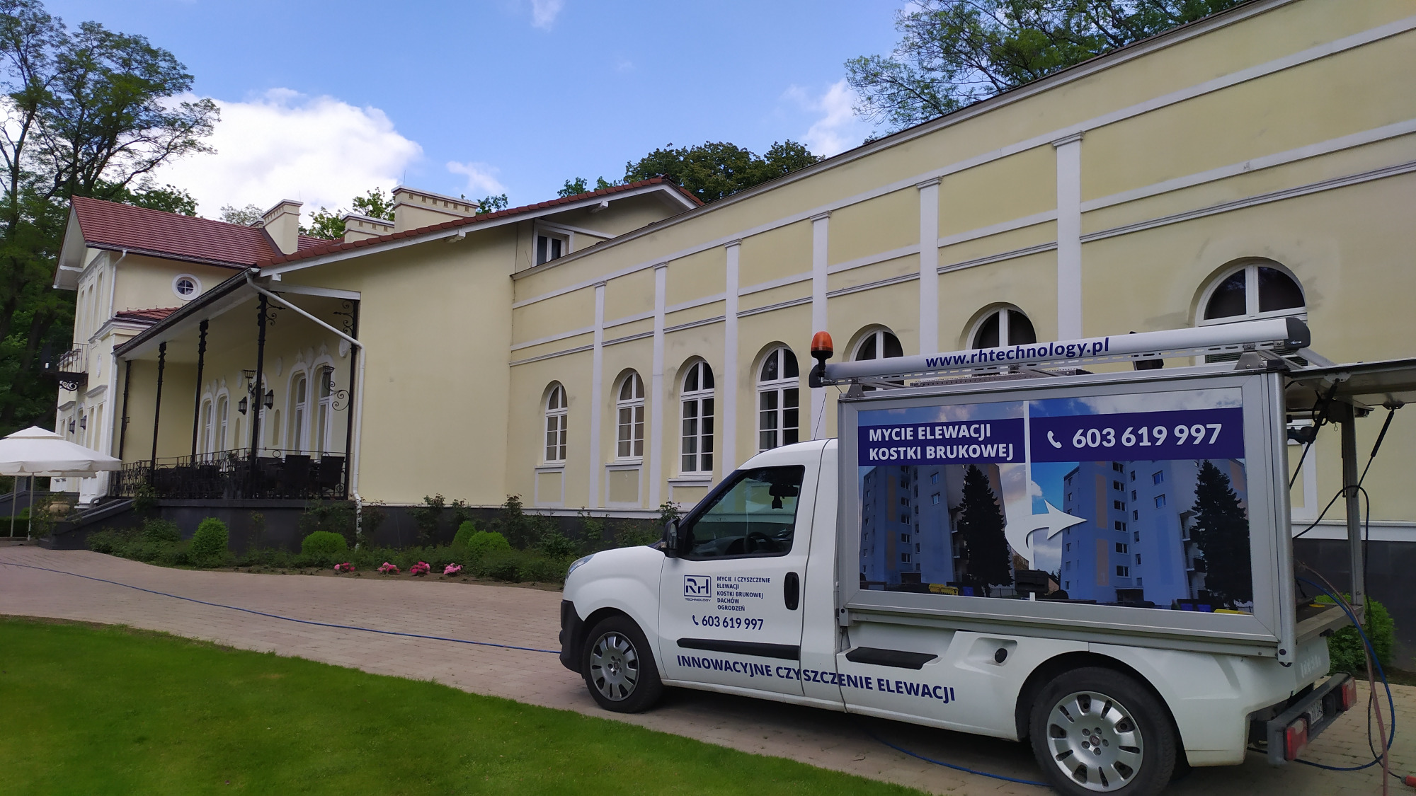 Biały samochód dostawczy z reklamą usług czyszczenia elewacji i kostki brukowej zaparkowany przed budynkiem o jasnej elewacji z rzędem łukowych okien.