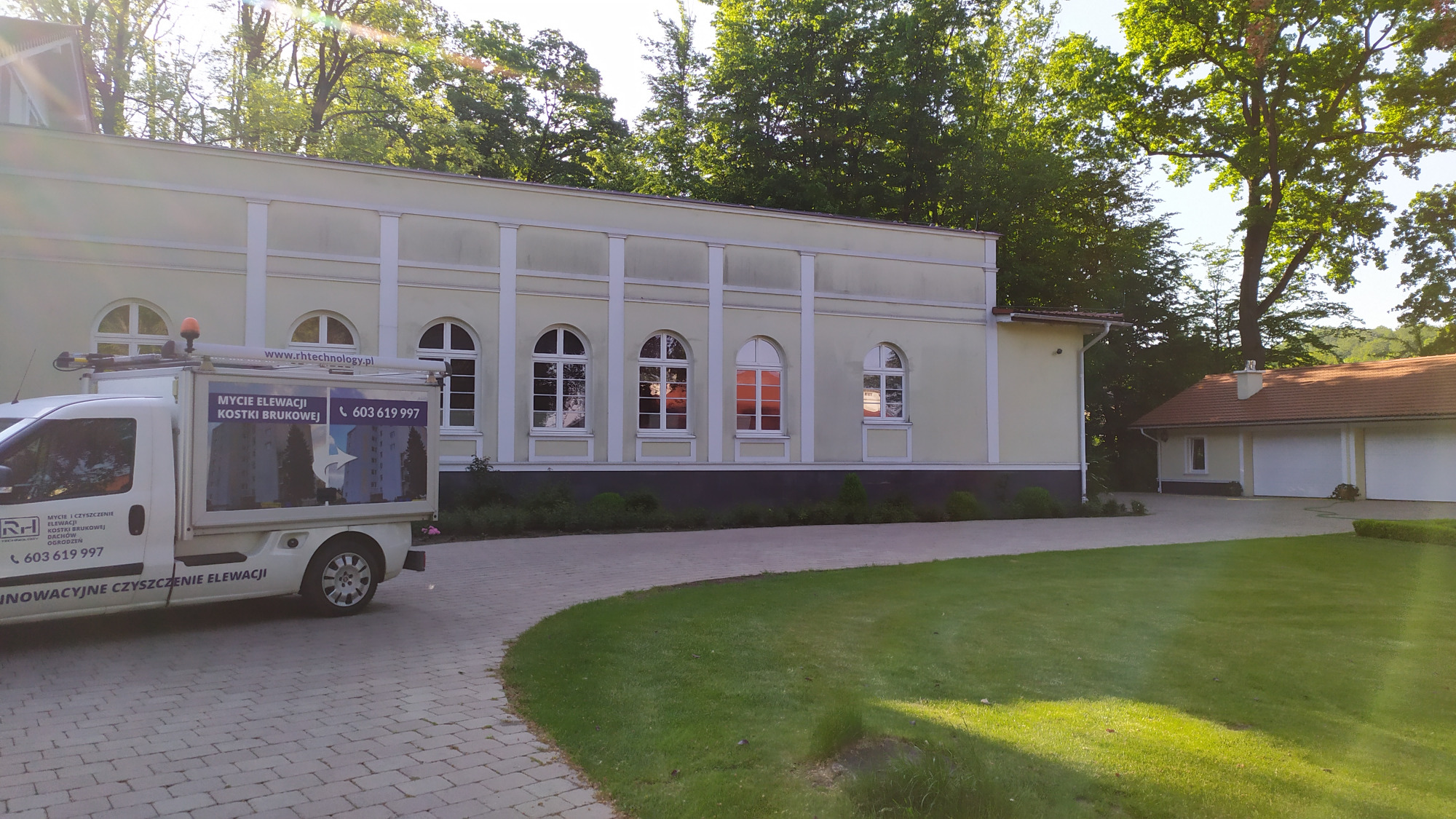 Jasna elewacja budynku z rzędami okien z białymi ramami, na pierwszym planie samochód dostawczy z reklamą mycia elewacji i kostki brukowej, otoczenie zielone.