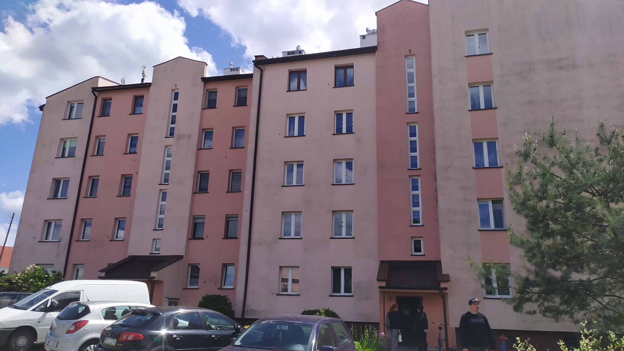 Wielorodzinny budynek mieszkalny o pastelowych elewacjach, z widocznymi zaciekami i zabrudzeniami, przed budynkiem zaparkowane samochody i osoby wchodzące do klatki.