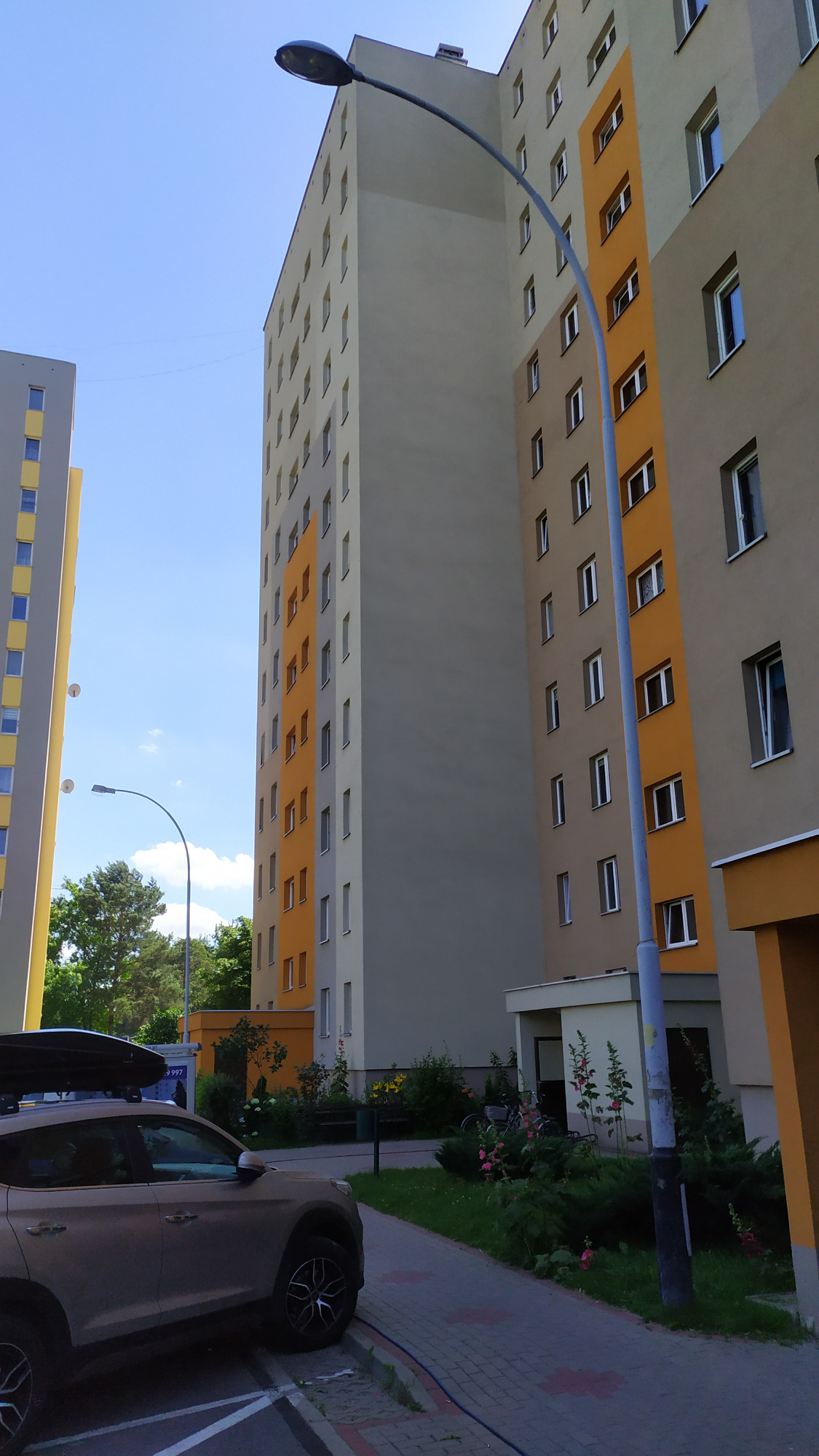 Wysoki blok mieszkalny z kolorową elewacją w odcieniach szarości i pomarańczy, widziany z dołu, z zaparkowanym szarym SUV-em z bagażnikiem dachowym na pierwszym planie.