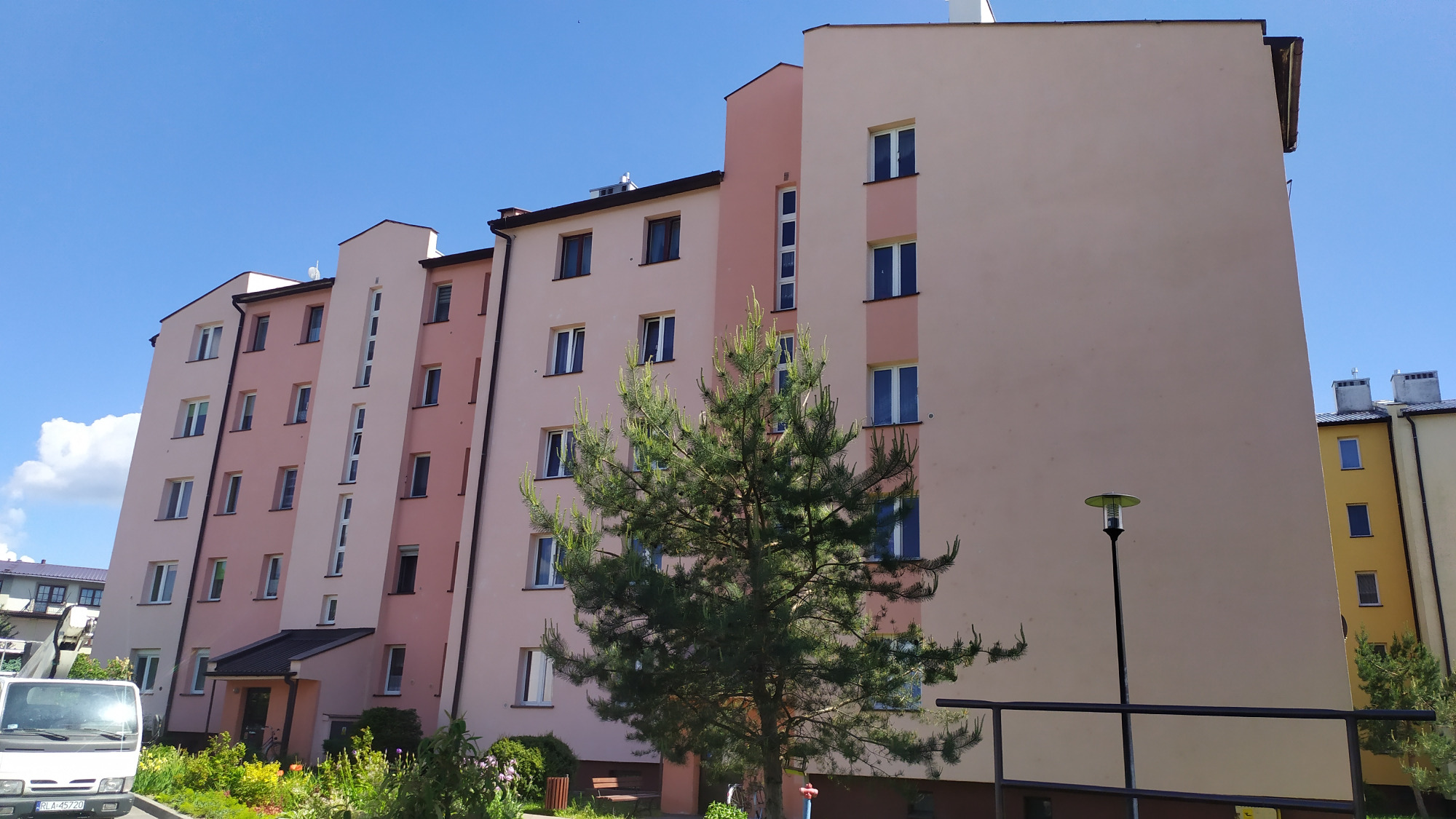 Wielopiętrowy budynek mieszkalny z elewacją w odcieniach różu, z widocznym fragmentem zaparkowanego białego samochodu dostawczego i drzewem iglastym na pierwszym planie.
