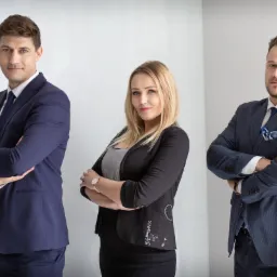 Trzy osoby w formalnych strojach stojące na tle białej ściany, kobieta w środku z ozdobnym wzorem na marynarce