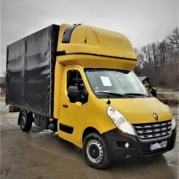 Żółty Renault Master z czarną plandeką na mokrym placu, widoczne drzewa w tle, tablica z ogłoszeniem na przedniej szybie.