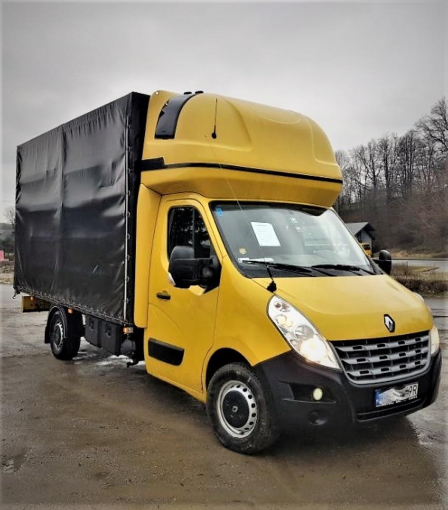 Żółty Renault Master z czarną plandeką na mokrym placu, widoczne drzewa w tle, tablica z ogłoszeniem na przedniej szybie.