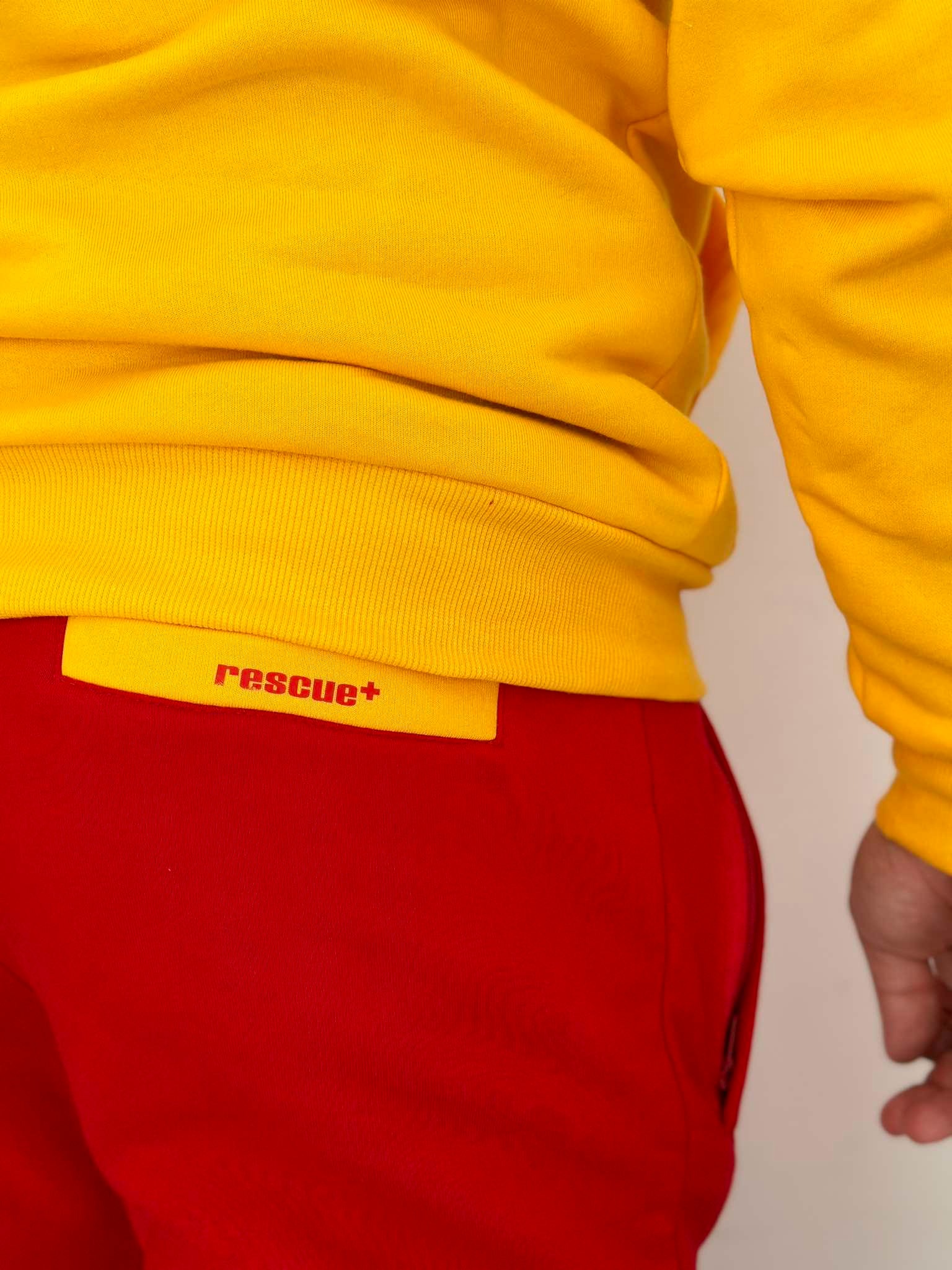 Fragment stroju ratownika: żółta bluza z napisem 'rescue+' i czerwone spodnie dresowe.
