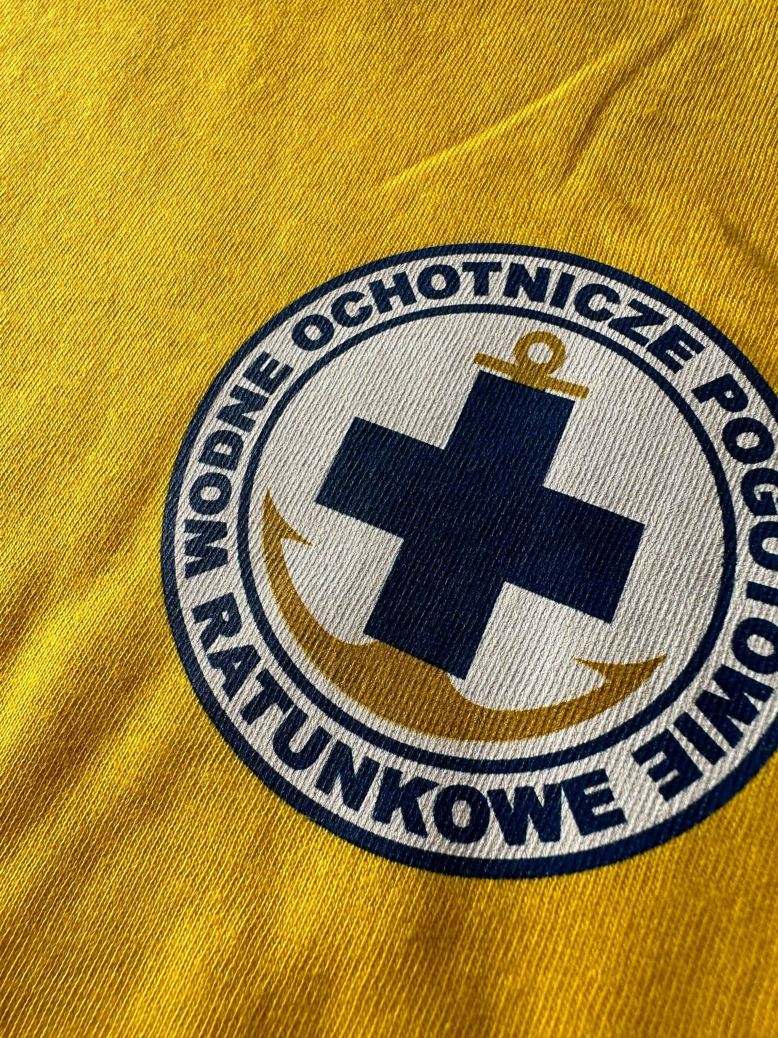 Żółta koszulka z nadrukowanym okrągłym logo Wodne Ochotnicze Pogotowie Ratunkowe z krzyżem i kotwicą.