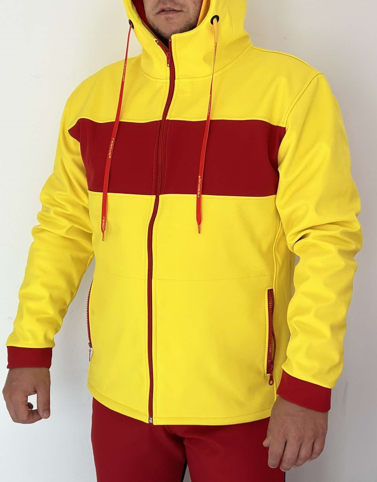 Mężczyzna prezentuje żółto-czerwoną bluzę z kapturem, z zamkiem błyskawicznym i czerwonymi sznurkami z napisem 'rescue+', widoczne spodnie w kolorze czerwonym.