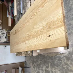 Blat z jasnego drewna o wyraźnym usłojeniu, widoczny w warsztacie stolarskim, z narzędziami na ścianie w tle.