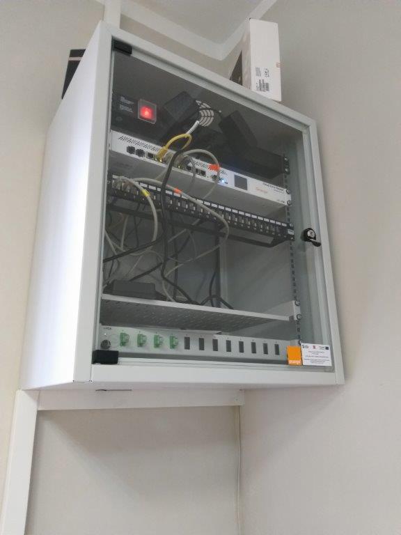 Biała szafa telekomunikacyjna z oszklonymi drzwiami, zamontowana na ścianie, widoczne kable, router i listwa zasilająca, pudełko na górze szafy.