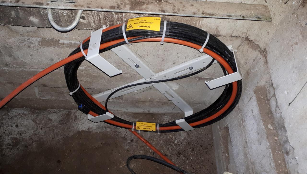 Kabel światłowodowy OKH10138 zwinięty w okrąg, przymocowany do sufitu piwnicy za pomocą białego, metalowego uchwytu krzyżowego.