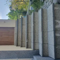 Zewnętrzne schody z szarej kostki prowadzące do drzwi wejściowych, widok z dołu, zieleń w tle, słoneczny dzień.