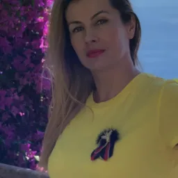 Portret kobiety z blond włosami w żółtej koszulce na tle kwitnącej bugenwilli i morza. Makijaż z czerwoną szminką. Na koszulce broszka w kształcie baletki.