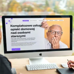 Dwie osoby przeglądają stronę internetową na komputerze iMac, prezentującą ofertę opieki domowej dla osób starszych, z zaskoczonym starszym panem na banerze.