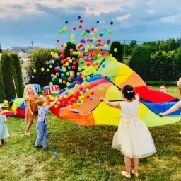 Grupa dzieci bawi się na trawie kolorową chustą animacyjną, podrzucając w górę mnóstwo kolorowych piłeczek. W tle widoczne drzewa i domy.