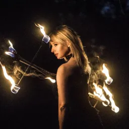Kobieta z blond włosami, w profilu, otoczona płonącymi pochodniami podczas pokazu fire show w ciemnościach.